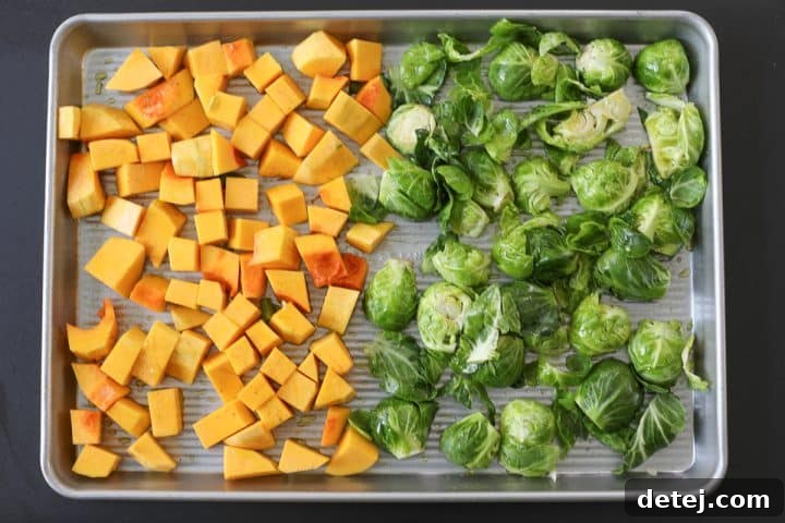 Raw butternut squash and Brussels sprouts seasoned with oil, salt, and pepper on a sheet tray before roasting.