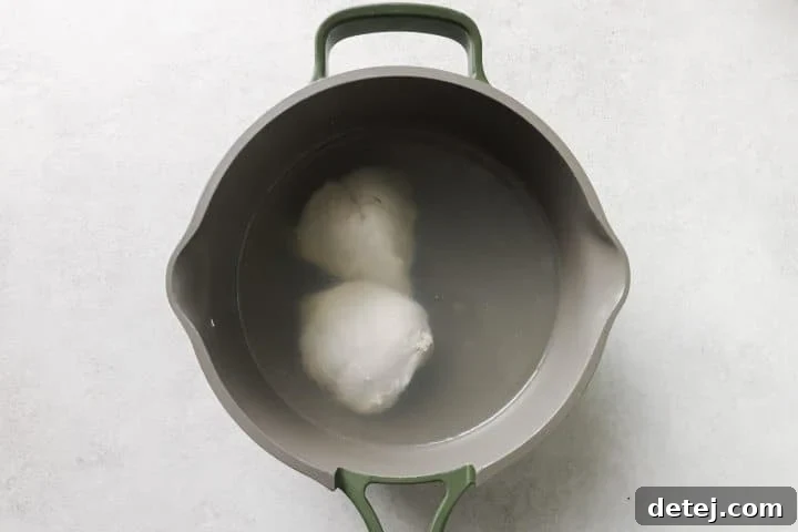 Close-up of chicken breasts simmering in a pot of clear broth on a stovetop, illustrating the boiling process.