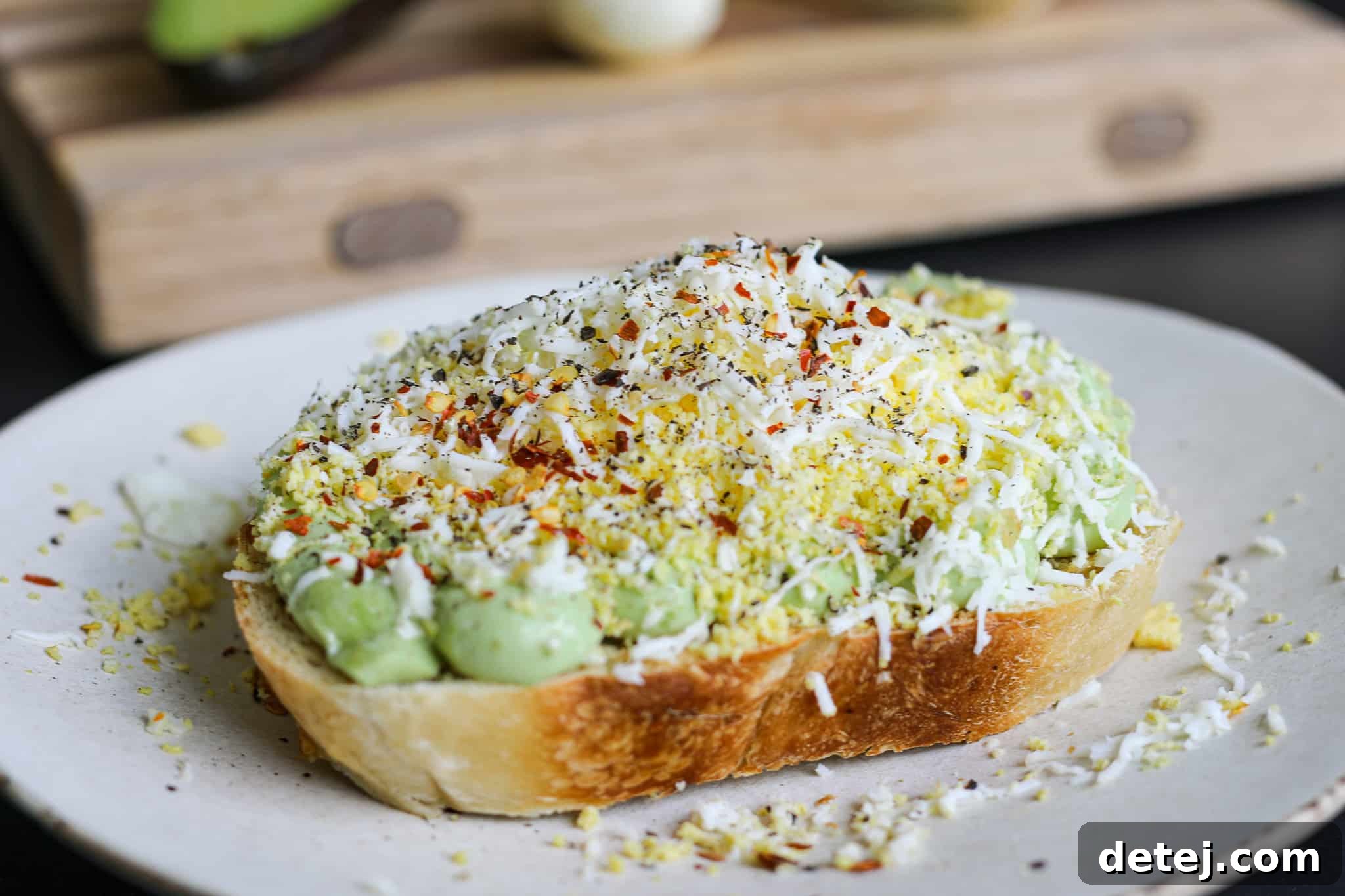 Close-up of the finished Grated Egg Avocado Toast with creamy mousse, fluffy grated egg, and red pepper flakes, ready to be served on a rustic wooden board.