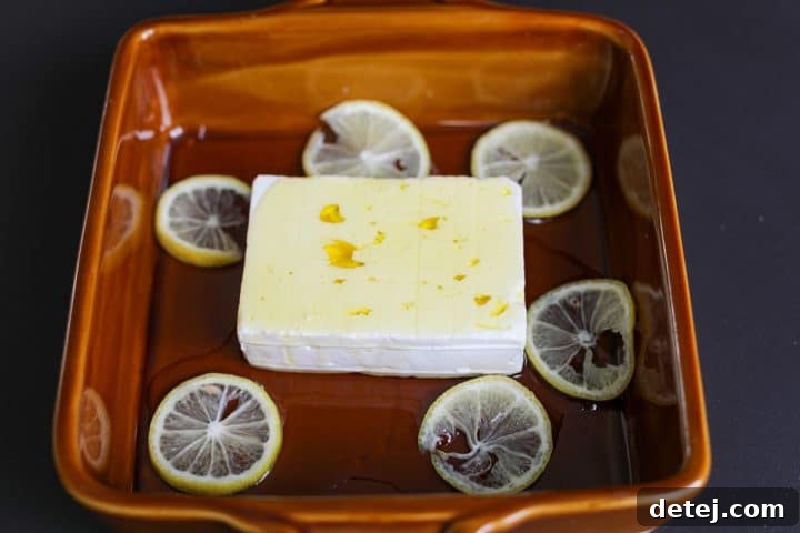 A block of feta in a ceramic baking dish, drizzled with olive oil and surrounded by seedless lemon slices.