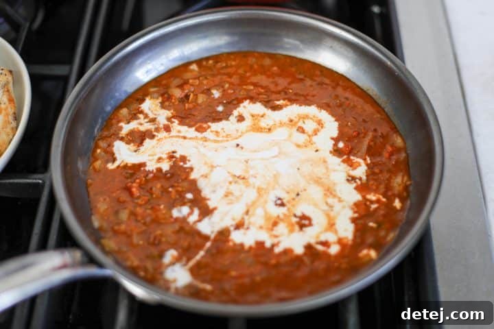 Heavy cream being added to the simmering tomato-pesto sauce, stirring to create a smooth, rich consistency.
