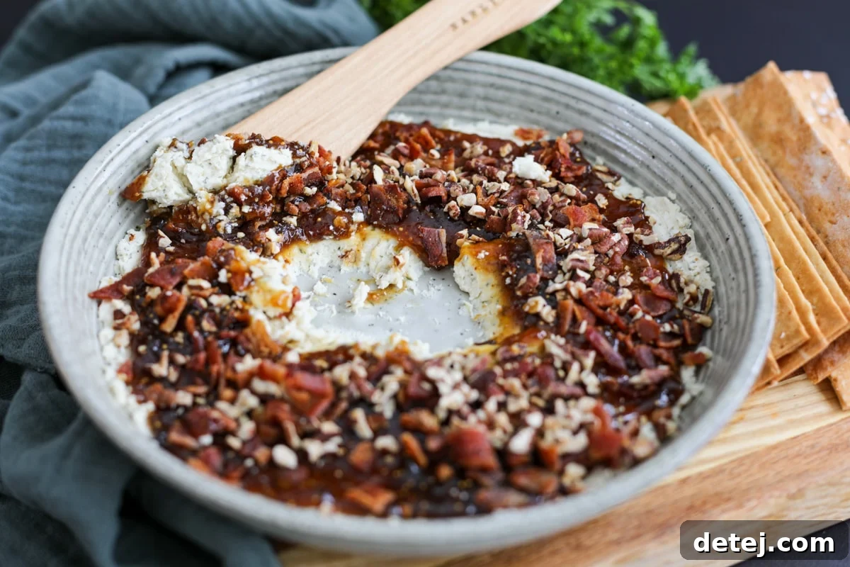 A serving of the Boursin cheese fig dip, with a cracker already dipped in, showing the layers of cheese, jam, bacon, and pecans.