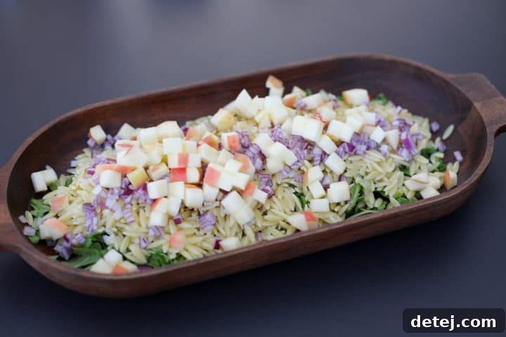 A close-up shot of the Chopped Fall Pasta Salad being assembled in a bowl, showing the orzo, arugula, apples, red onion, and bacon.