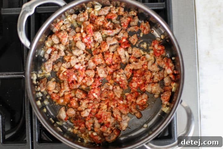 Tomato paste being stirred into the browned sausage and sautéed aromatics in a hot pan.