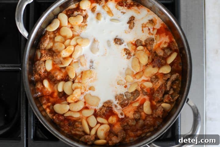Heavy cream being poured into the mixture of sausage, beans, and aromatics in a pan.