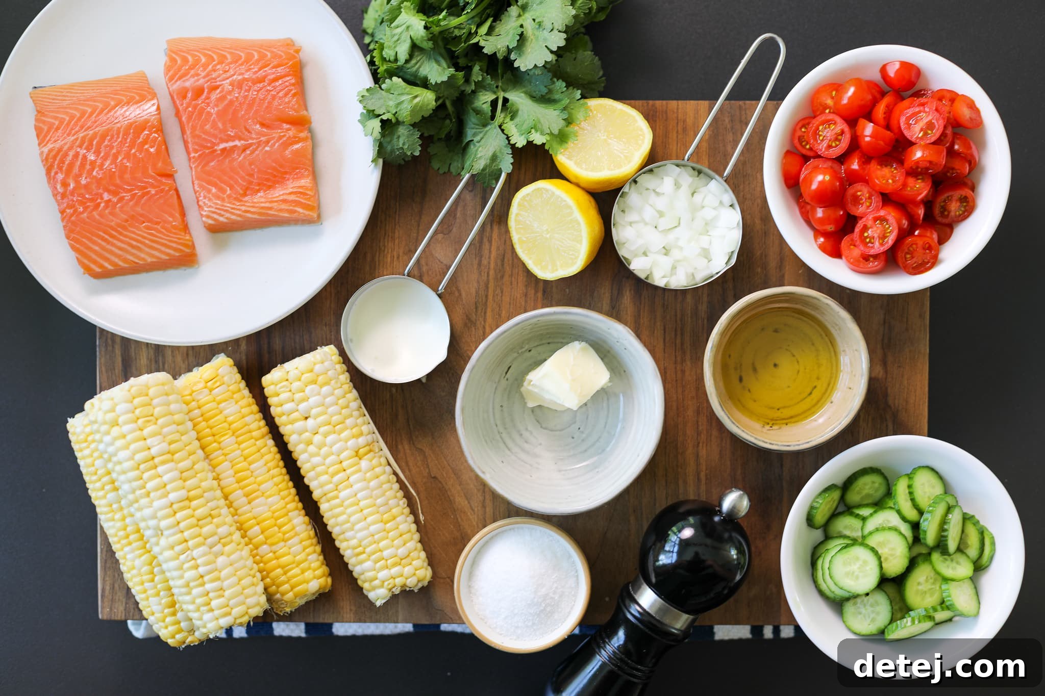 A collection of fresh ingredients laid out on a wooden board, including salmon fillets, corn cobs, cherry tomatoes, cucumber, onion, and herbs, ready for the grilled salmon, corn puree, and tomato salad recipe.