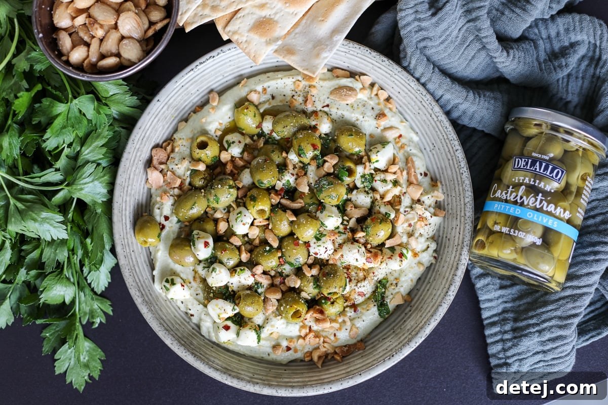A vibrant bowl of marinated olives and mozzarella over creamy whipped pesto ricotta, garnished with fresh herbs and chopped almonds.
