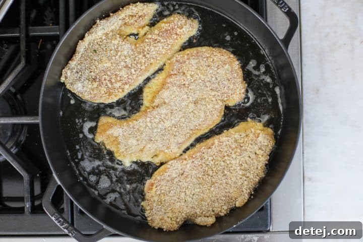 Two breaded chicken cutlets shallow frying in a pan, achieving a golden-brown crust.